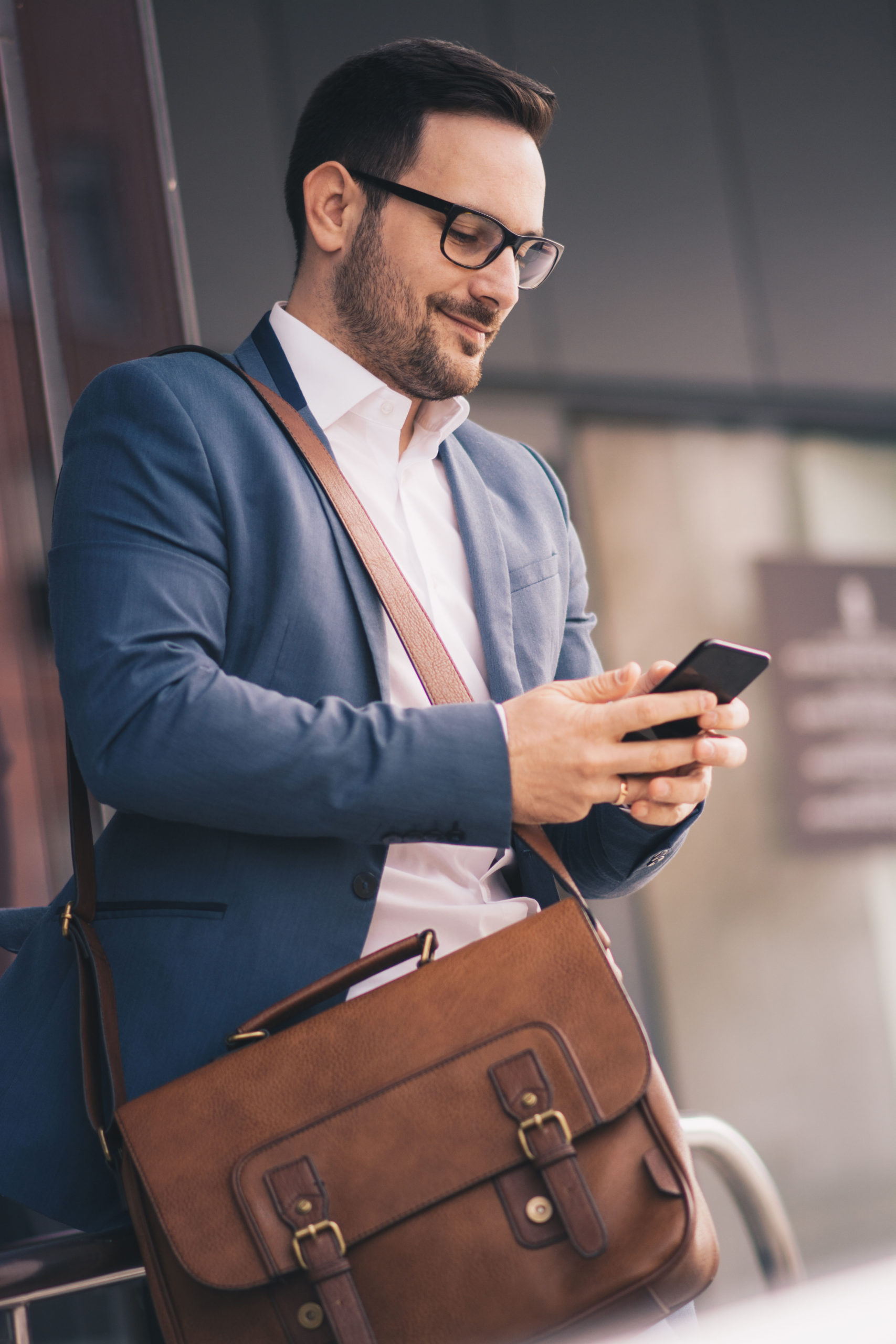  A young businessman outdoors, looking at his phone. He is carrying a bag over his shoulder. 